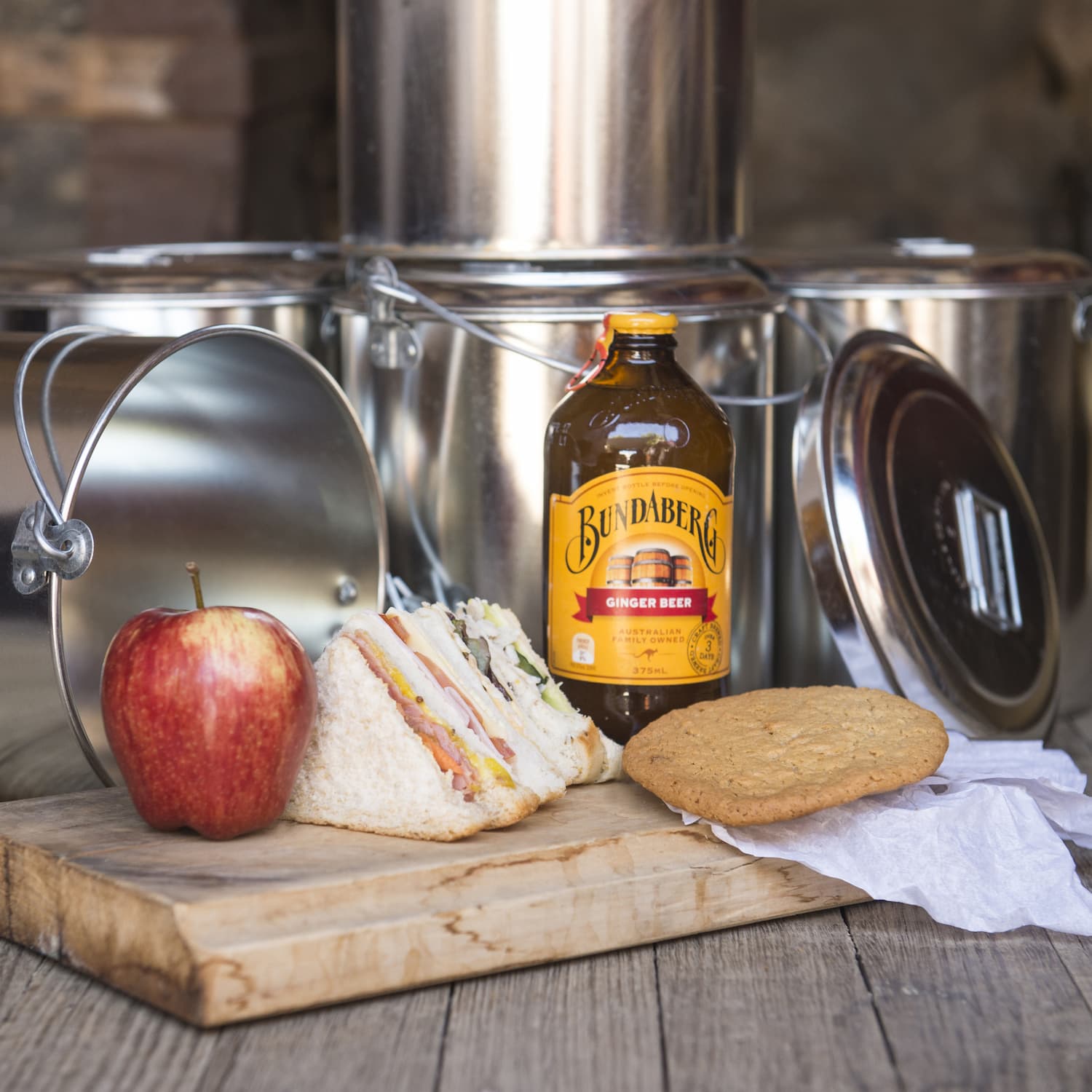 A billy-can lunch including a cut sandwich, anzac biscuit, apple, and ginger beer, presented on a timber board