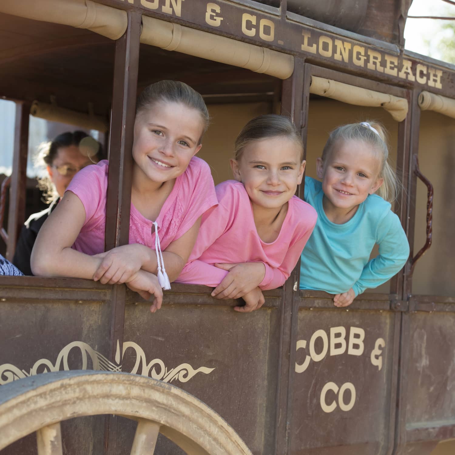 Three young girls smiling and leaning out the open window of a Cobb and Co Stagecoach