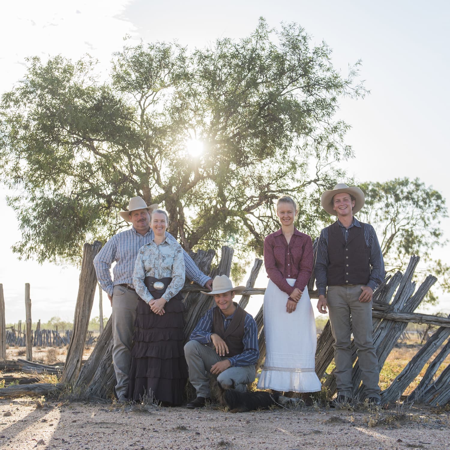 The Kinnon family standing in front of old wooden fence palings on Nogo Station. Tree behind with sun shining through