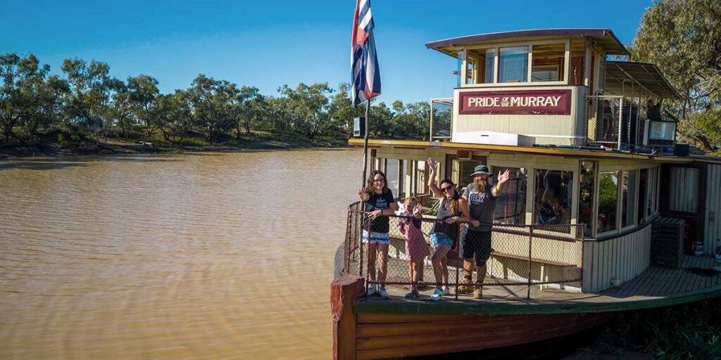 Pride of the Murray paddlewheeler anchored alongside the riverbank. A family of four waving from the front deck.
